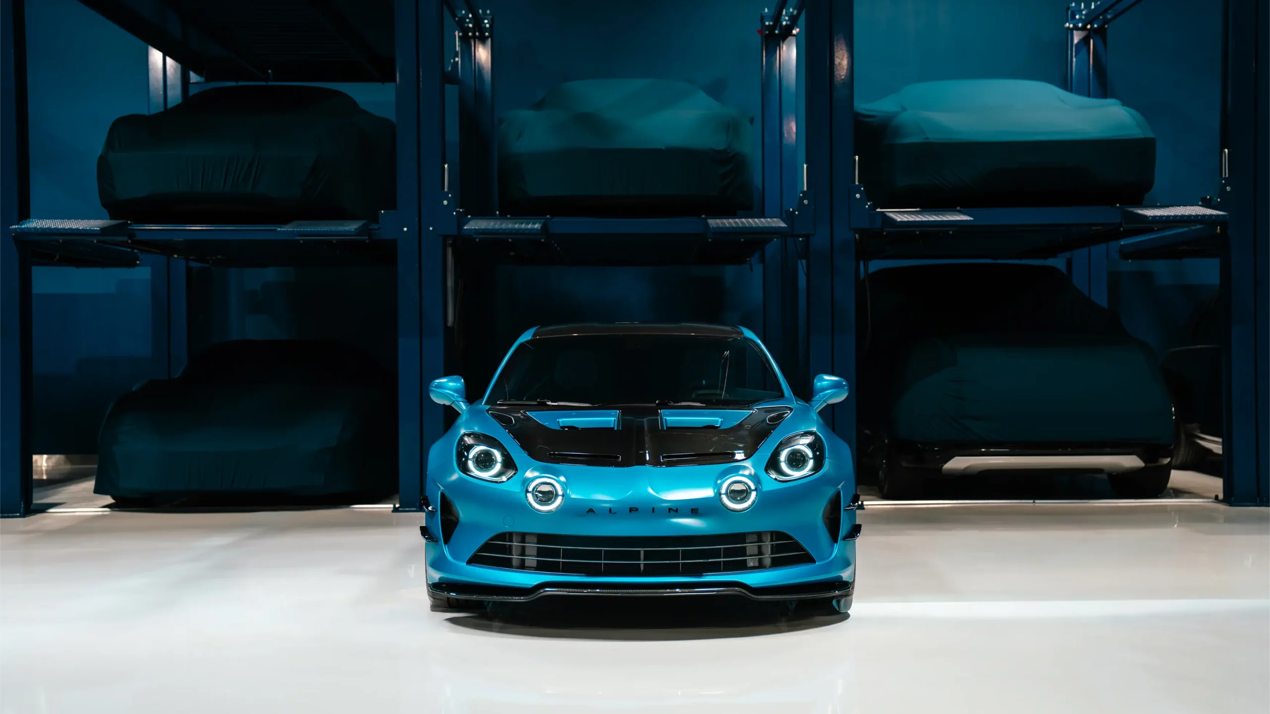 A blue Alpine A110 photographed head-on under low light, parked in front of a two-tier steel storage rack holding cars resting beneath fitted covers inside The Collectors Hangar.