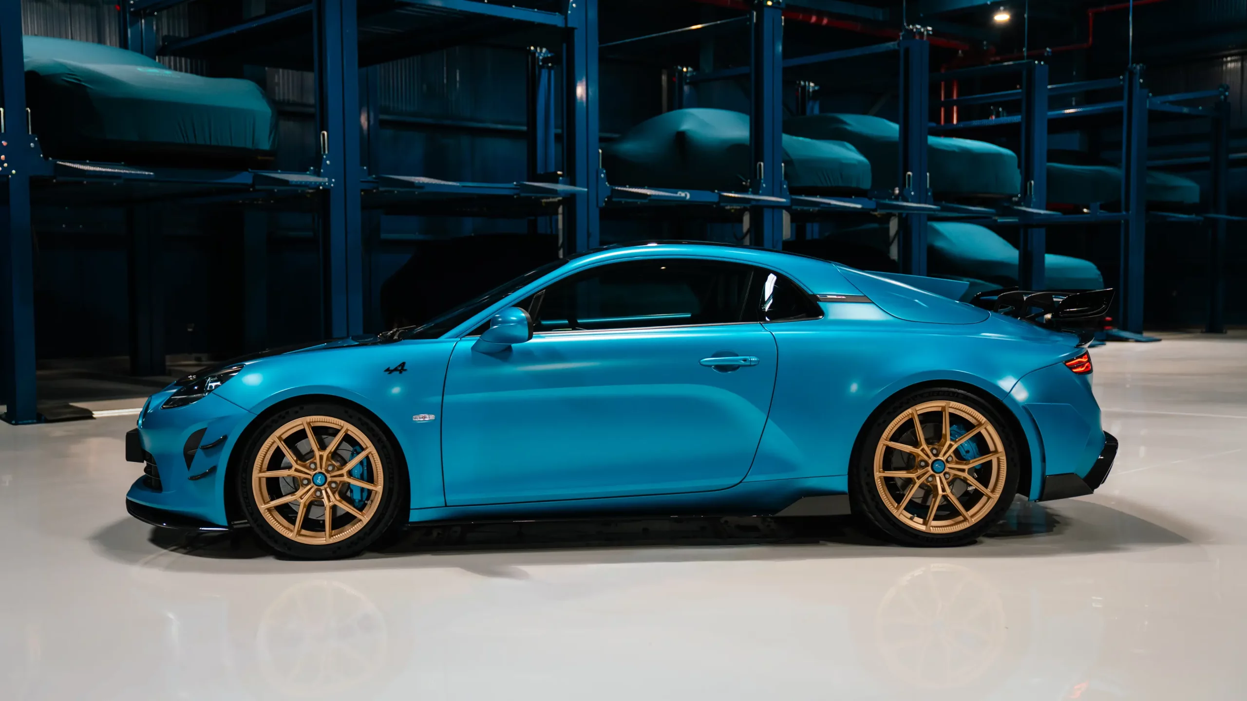 An Alpine A110 in profile on the polished floor of The Collectors Hangar, with two tiers of cars resting under fitted covers on the storage racks behind it