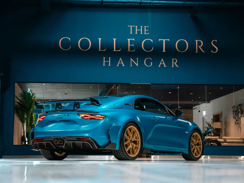 A light-blue Alpine A110 R seen from the rear three-quarter, its tall carbon rear wing and quad centre exhausts catching the light, parked on a reflective floor beneath the gold THE COLLECTORS HANGAR lettering set on the deep blue wall, with the glass-fronted lounge lit behind it.