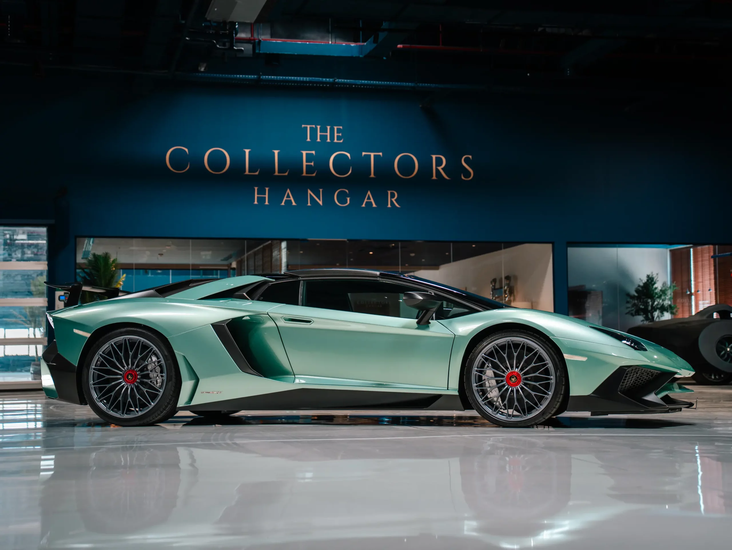 A mint-green Lamborghini Aventador SV seen in profile on a mirror-polished floor, standing beneath the illuminated gold THE COLLECTORS HANGAR lettering on the facility's dark navy wall.