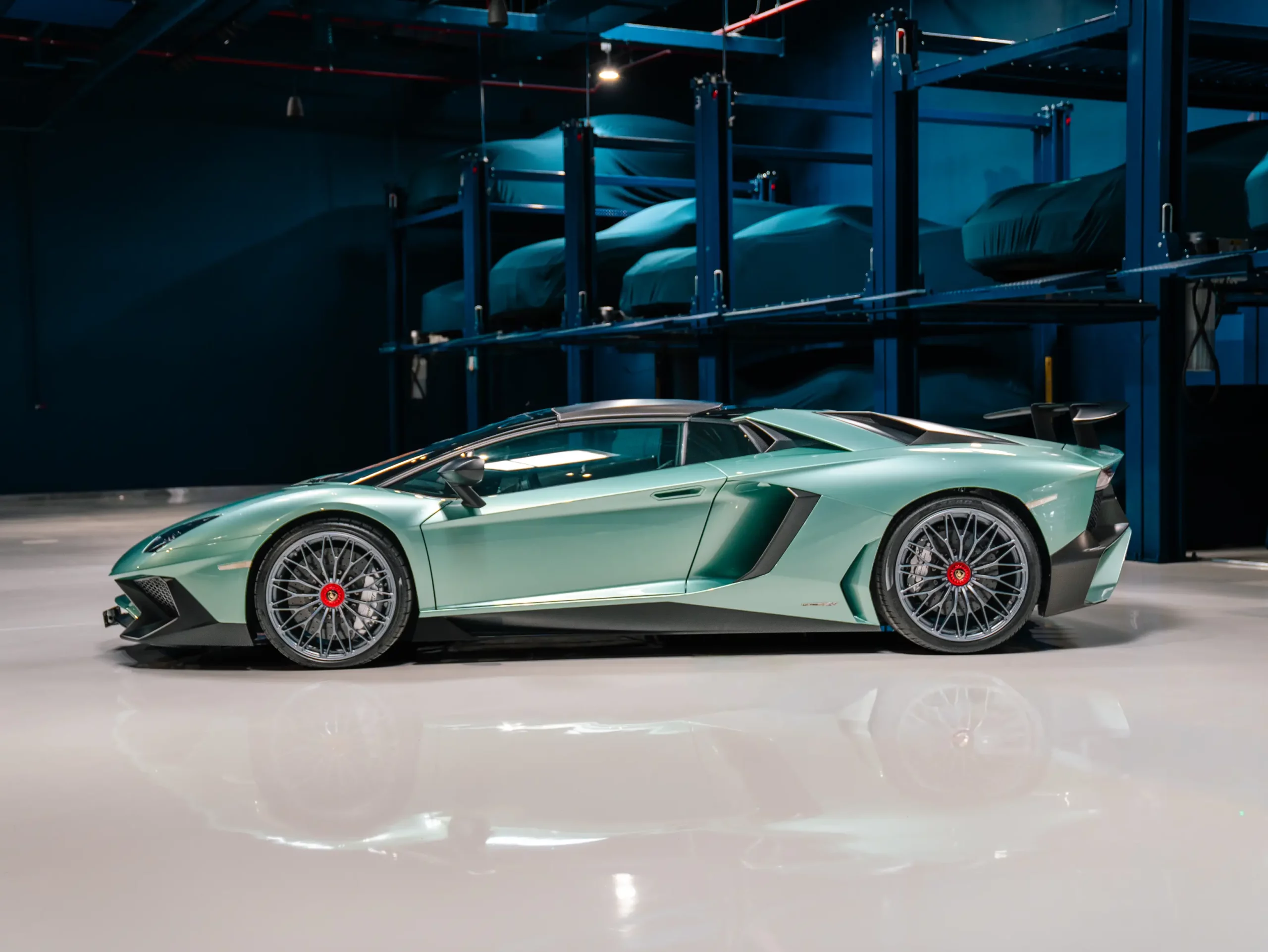 A mint-green Lamborghini Aventador SV in full profile on a reflective floor, with two levels of steel storage racking holding cars under fitted covers in the darkness behind it.