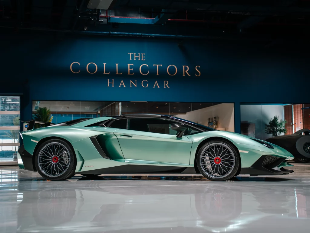 A mint-green Lamborghini Aventador SV in full profile on a mirror-polished floor, standing beneath the illuminated gold THE COLLECTORS HANGAR lettering on the facility's deep navy wall.