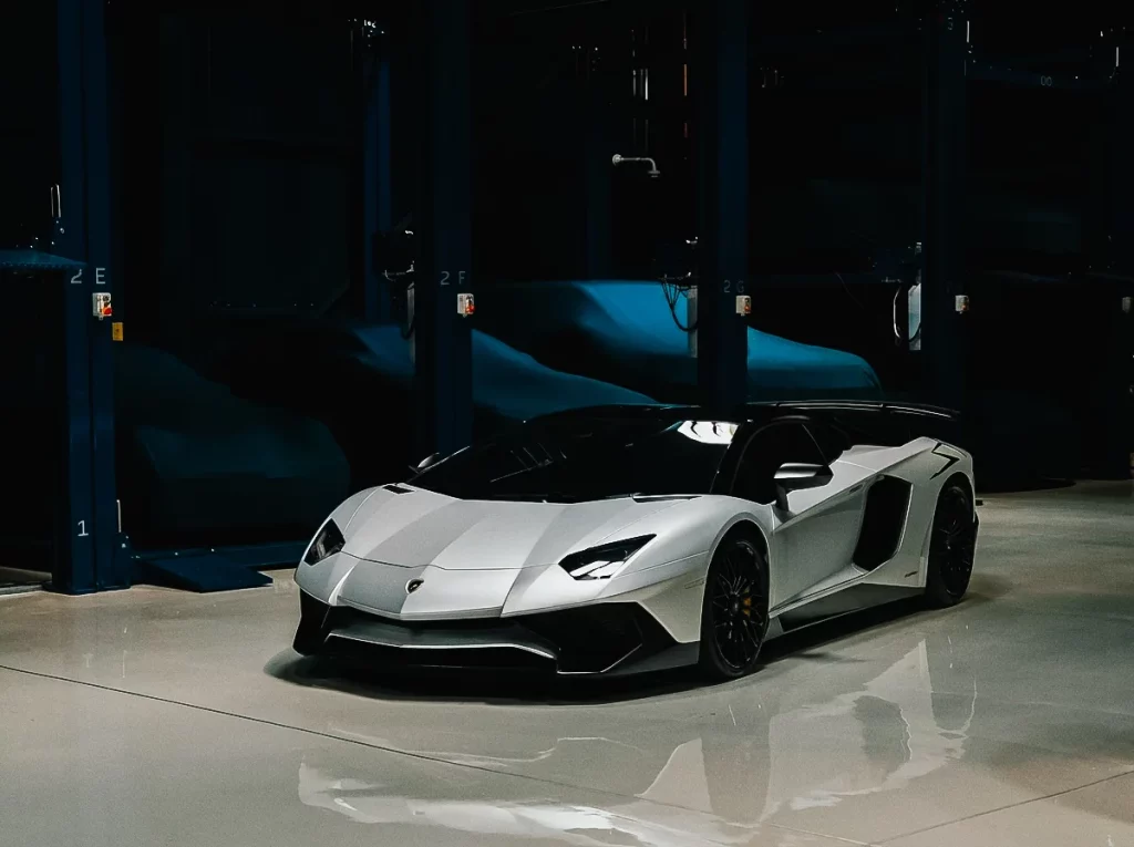 A white Lamborghini Aventador SV lit from above in a darkened storage bay at The Collectors Hangar, its reflection on the pale polished floor, with covered cars resting on the blue racking behind it.