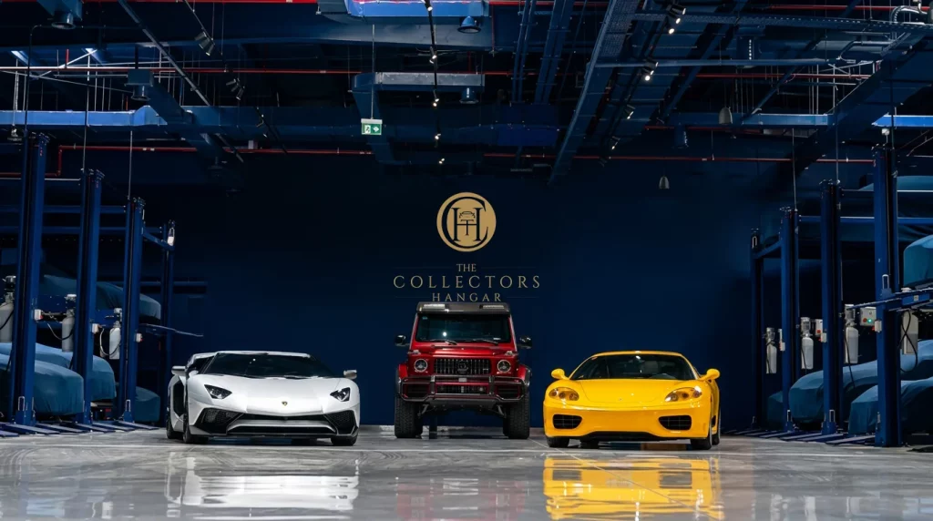 Three collector cars lined up head-on beneath the gold Collectors Hangar monogram and lettering on the deep blue end wall: a white Lamborghini Aventador SV on the left, a red Mercedes-AMG G-Class in the centre and a yellow Ferrari 360 on the right, mirrored in the polished floor with covered cars on the racks along both side walls.
