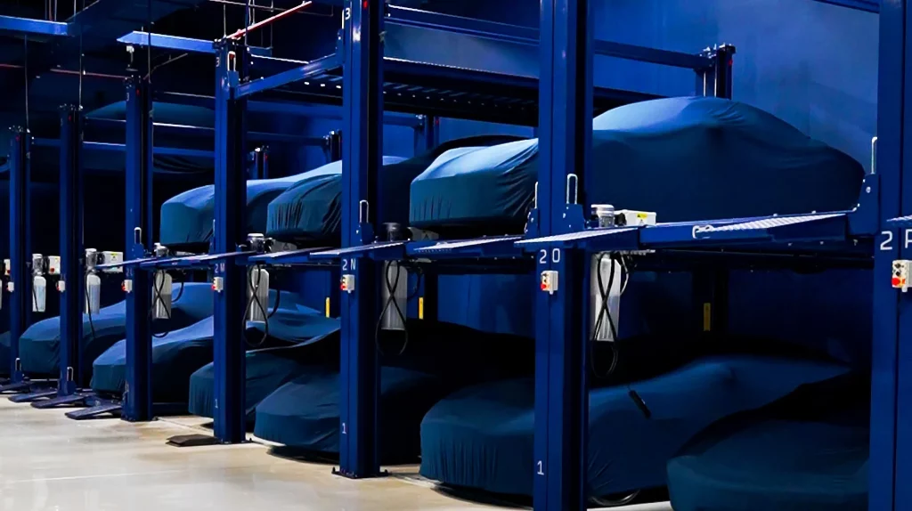 Rows of blue two-tier storage racks inside The Collectors Hangar, each bay holding a car beneath a fitted teal cover, with lettered and numbered bay markers on the posts and charging cables coiled beside them.