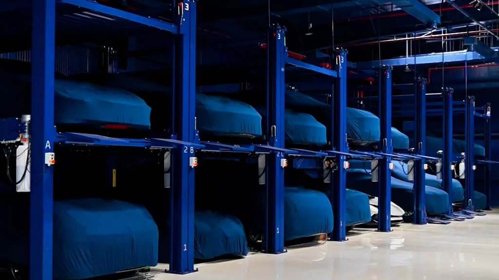 A long row of blue four-post storage racks receding down the hangar floor, every upper and lower bay filled with a car under a fitted teal cover, lettered and numbered bay markers on the posts and a pale polished floor running the length of the aisle.
